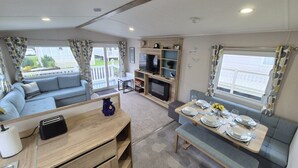 A cozy living area with a sofa, a dining table set for four, a TV, and a bookshelf. - Whitstable Neptune - Alberta Holiday Park (Whitstable)