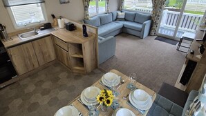 A compact living space with a dining area set for four, a kitchenette, a sofa, and a view of the outdoors. - Whitstable Neptune - Alberta Holiday Park (Whitstable)