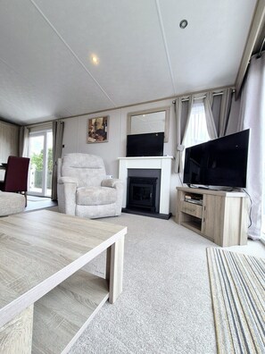 A modern living room with a sofa, a television, a fireplace, and a dining table. - The Helmsley Lodge - Alberta Holiday Park (Whitstable)