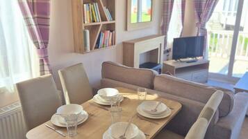 A dining area with a wooden table set for four, surrounded by cushioned chairs. A bookshelf with various books is visible in the background.