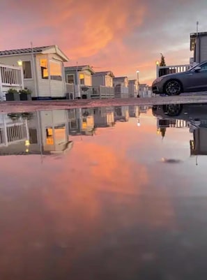 A row of white mobile homes with a car on a trailer in the background, reflecting on a calm water surface. - Whitstable’s Tranquility (Whitstable)