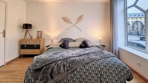 Un dormitorio con una cama grande, una mesita de noche de madera, una lámpara de pared y una ventana con vista a la calle.