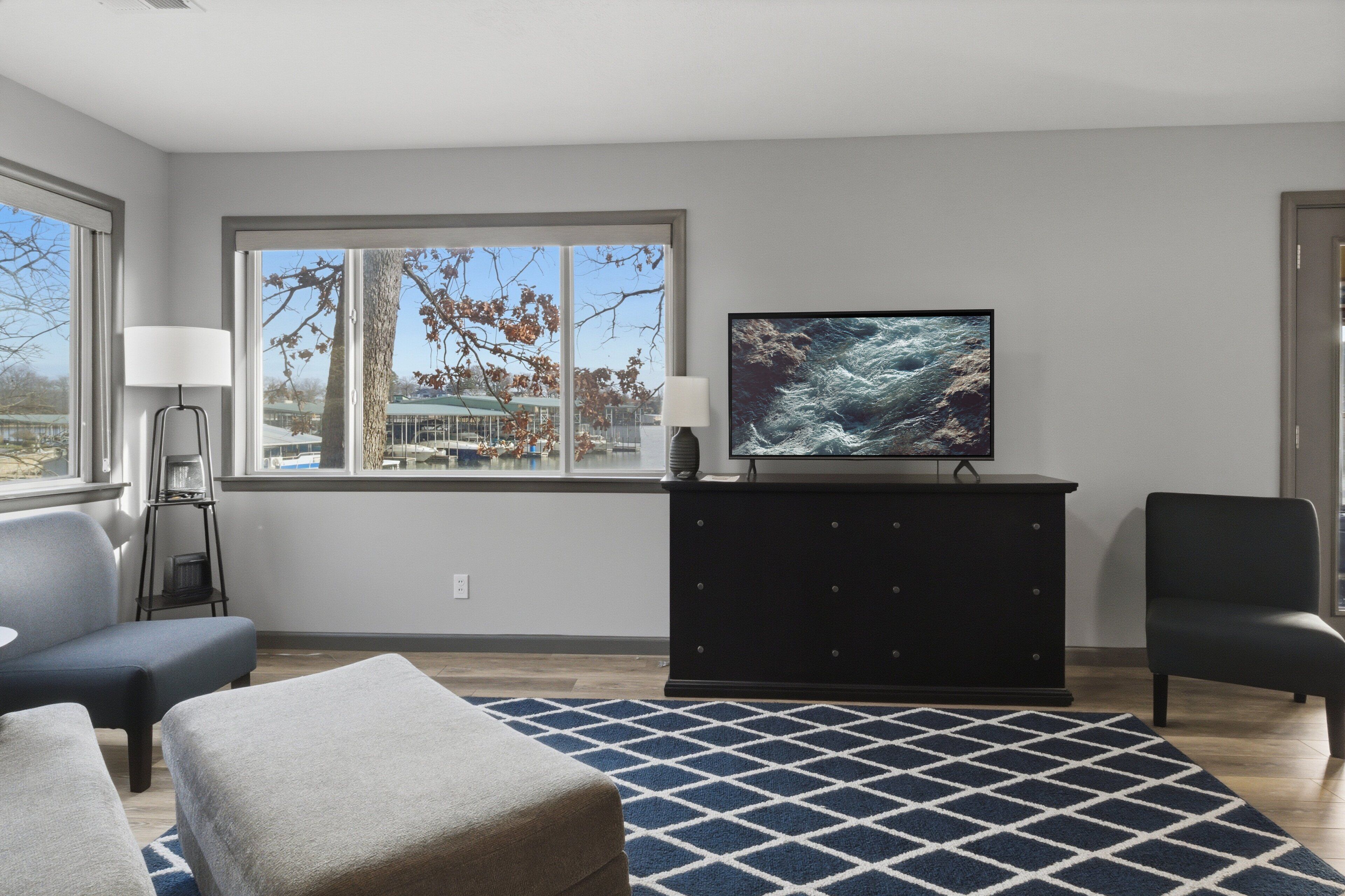 A modern living room with a large window, a flat-screen TV, a dark-colored dresser, and two armchairs.