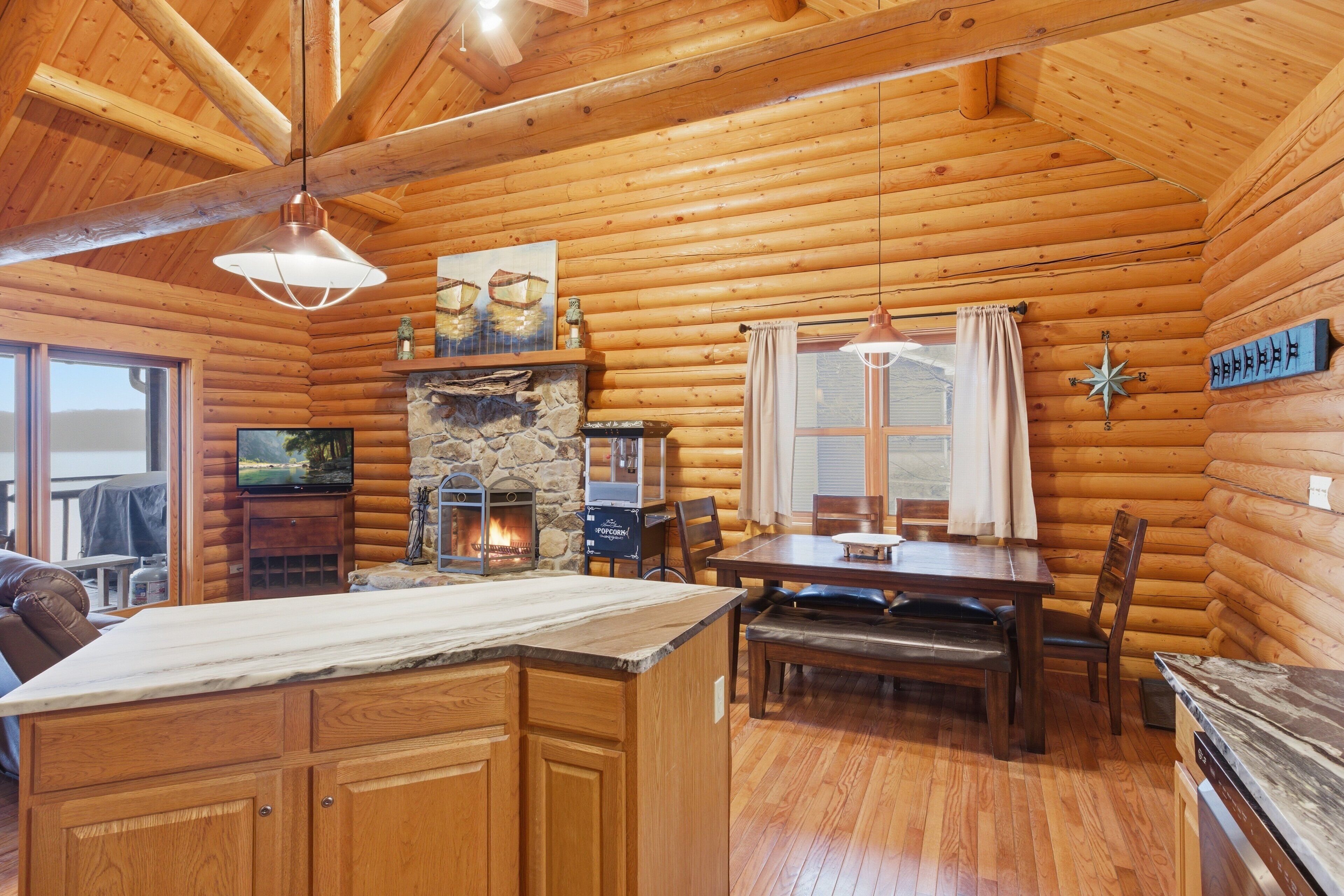 A log cabin interior with a stone fireplace, wooden dining table, and a large window with curtains.