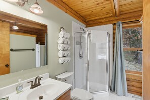 A bathroom with a wooden ceiling, a glass shower enclosure, a white sink, and a toilet. - Lakefront Log Cabin With Private Dock, Two Decks & Cozy Comfort at 35mm in Sunrise Beach (Sunrise Beach)