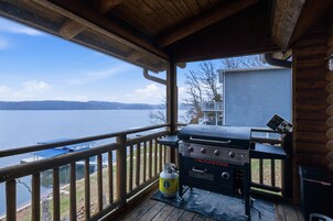 A wooden deck with a grill and propane tank, overlooking a lake and distant mountains.