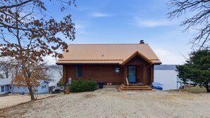 A wooden cabin with a metal roof, a front porch, and a gravel driveway. - Lakefront Log Cabin With Private Dock, Two Decks & Cozy Comfort at 35mm in Sunrise Beach (Sunrise Beach)