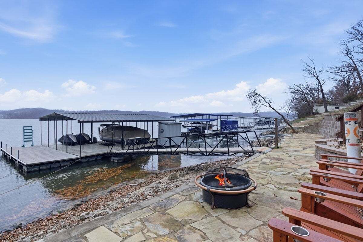 Ein Steg am Seeufer mit Feuerstelle, Holzstühlen und einer überdachten Sitzgruppe mit Tisch und Stühlen.