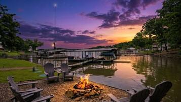 Un paisaje junto al lago con una fogata, sillas Adirondack, un muelle y un edificio con galería cubierta.