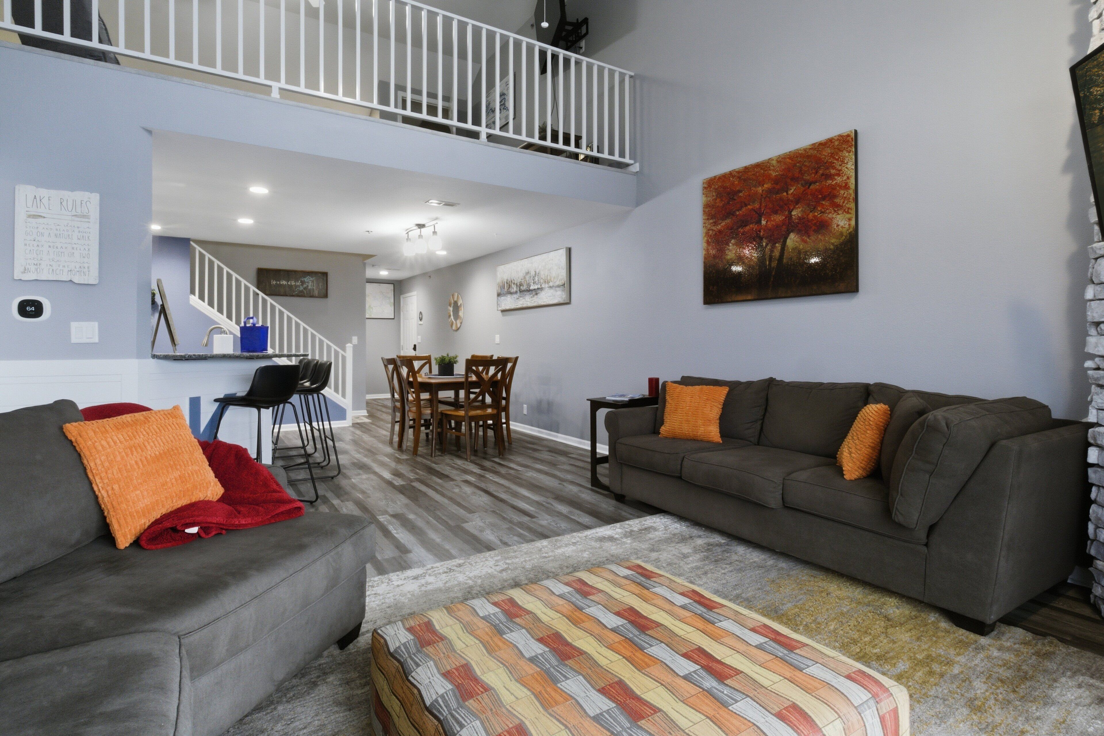 Una sala de estar con un sofá gris, un otomano estampado, una zona de comedor con mesa y sillas, y una escalera que conduce a un nivel superior.