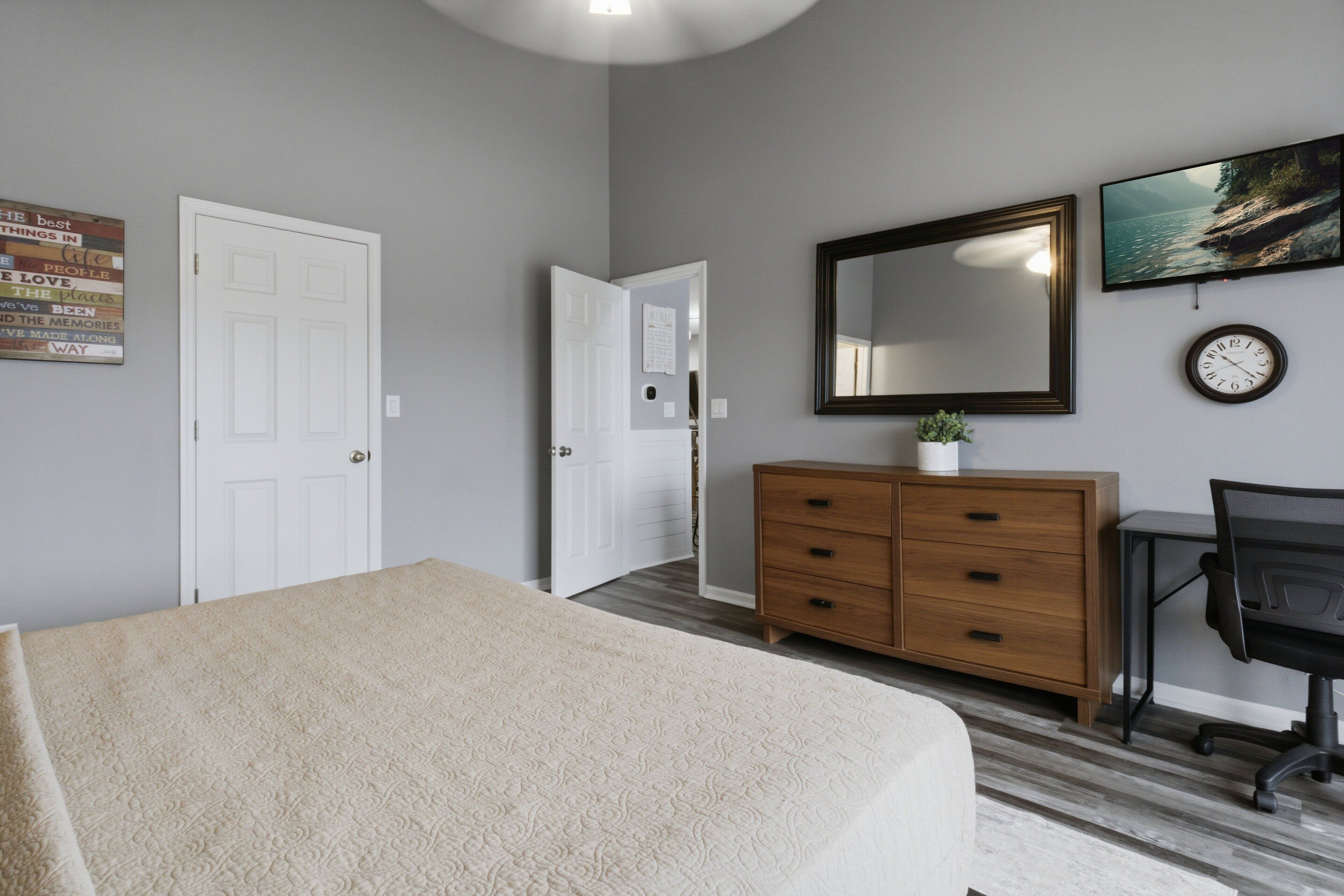 Un dormitorio con cama, cómoda de madera, escritorio, silla, espejo y un reloj en la pared.