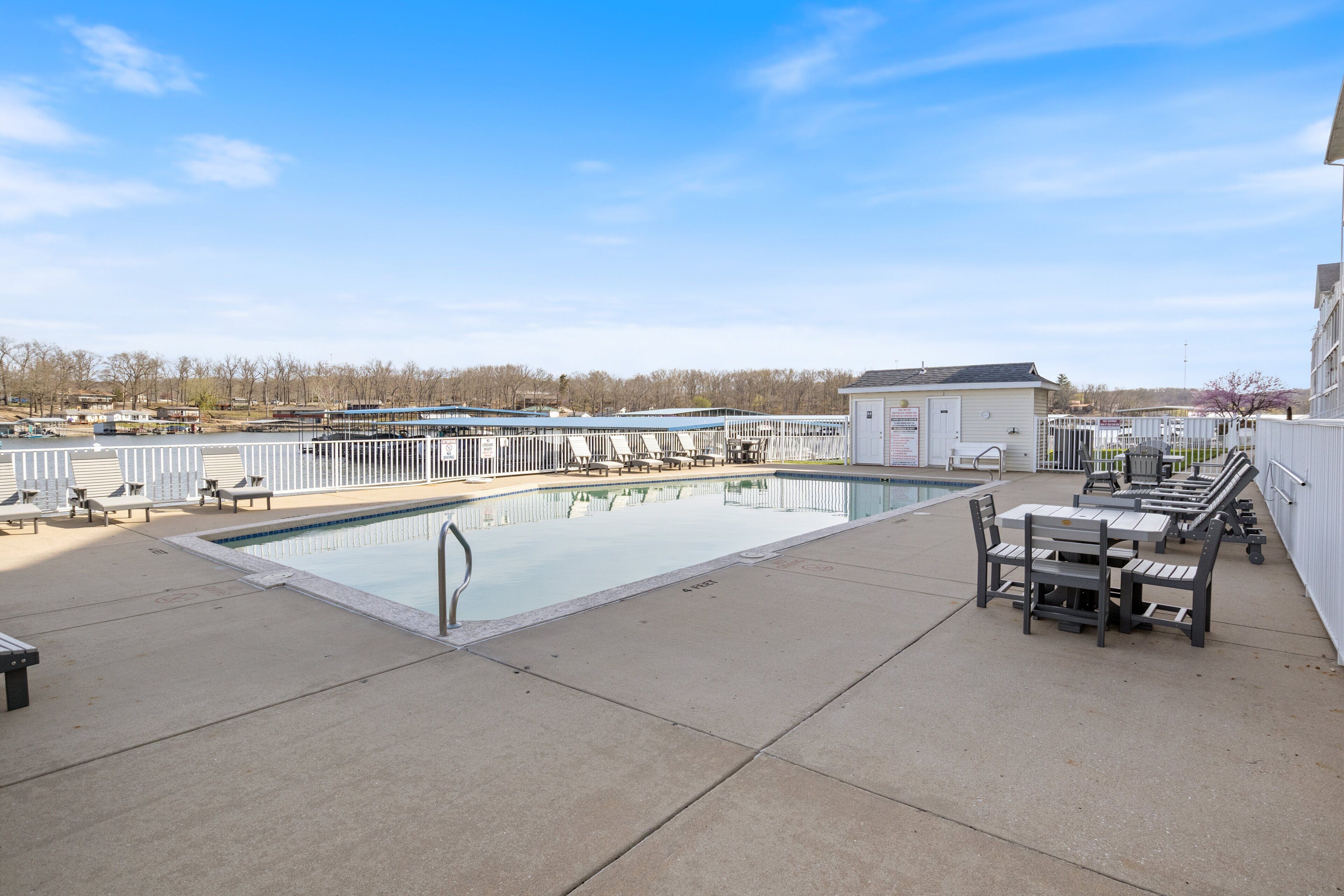 A swimming pool with lounge chairs and a table, surrounded by a fence and buildings in the background.