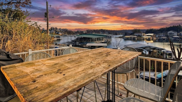 Una mesa de madera y sillas de metal en una terraza al aire libre, con vista a un río y edificios a lo lejos.