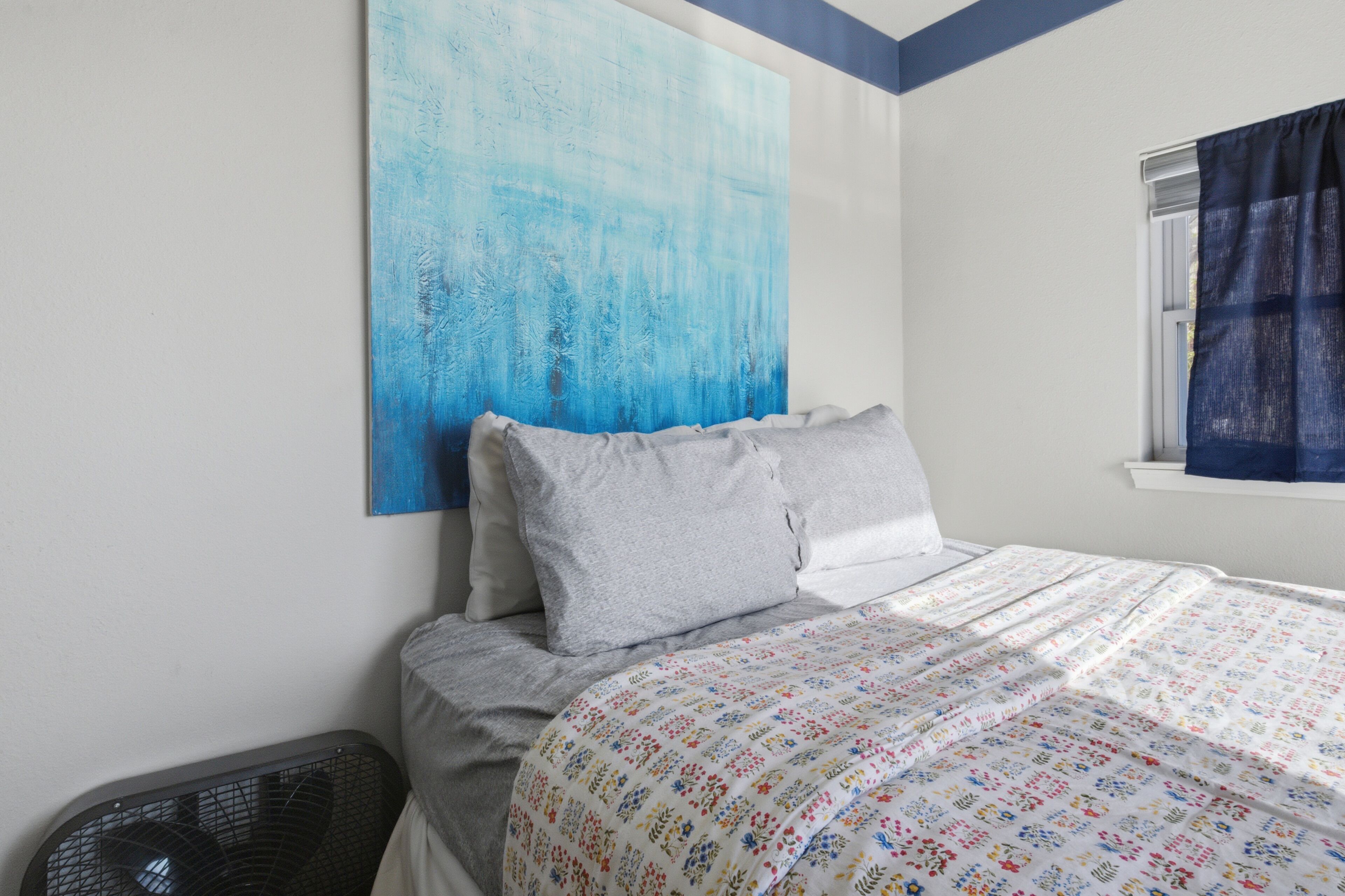 Een slaapkamer met een bed, kussens, een kunstwerk aan de muur, een raam met gordijnen en een vloerventilator.