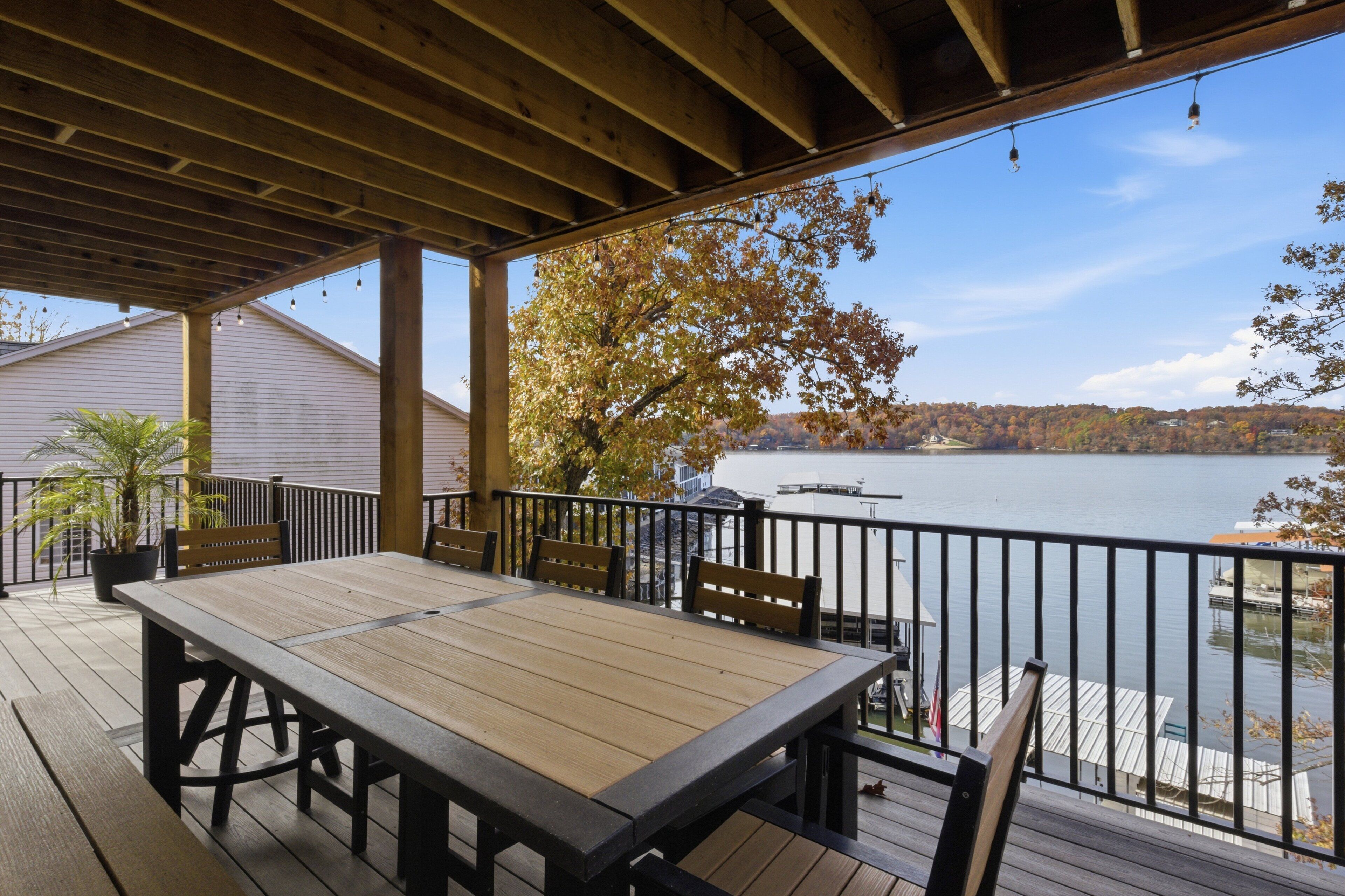 Een houten terras met een tafel en stoelen, met uitzicht op een rustig meer met boten en een pier, omringd door bomen met herfstkleuren.