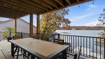 Een houten terras met een tafel en stoelen, met uitzicht op een rustig meer met boten en een pier, omringd door bomen met herfstkleuren.