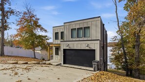A modern two-story house with a garage, surrounded by trees and a clear sky. - New Lakefront Home With Private Dock, Game Room & Firepit at 9MM (Lake Ozark)