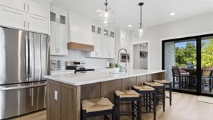 A modern kitchen with white cabinets, stainless steel appliances, a large island with bar stools, and a view through a glass door to the outdoors. - Modern Lakefront Escape The Twelve (Camdenton)