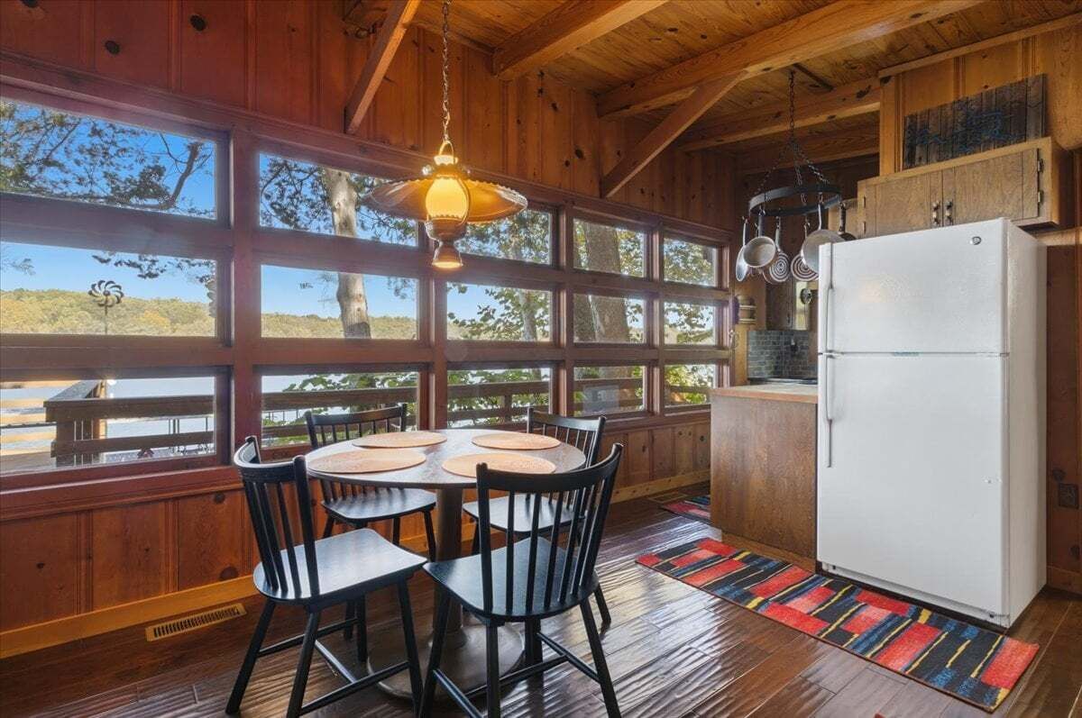A wooden interior with a dining area, large windows, and a refrigerator.