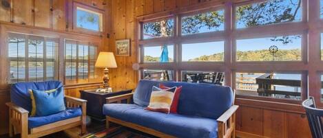 Interior de una cabaña de madera con un sofá azul, dos sillones, un tapete a rayas y grandes ventanales que ofrecen vistas al exterior.
