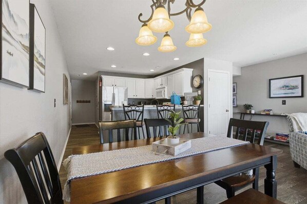 A dining area with a wooden table, chairs, and a chandelier. A kitchen is visible in the background. - Serenity Now Retreat (Camdenton)