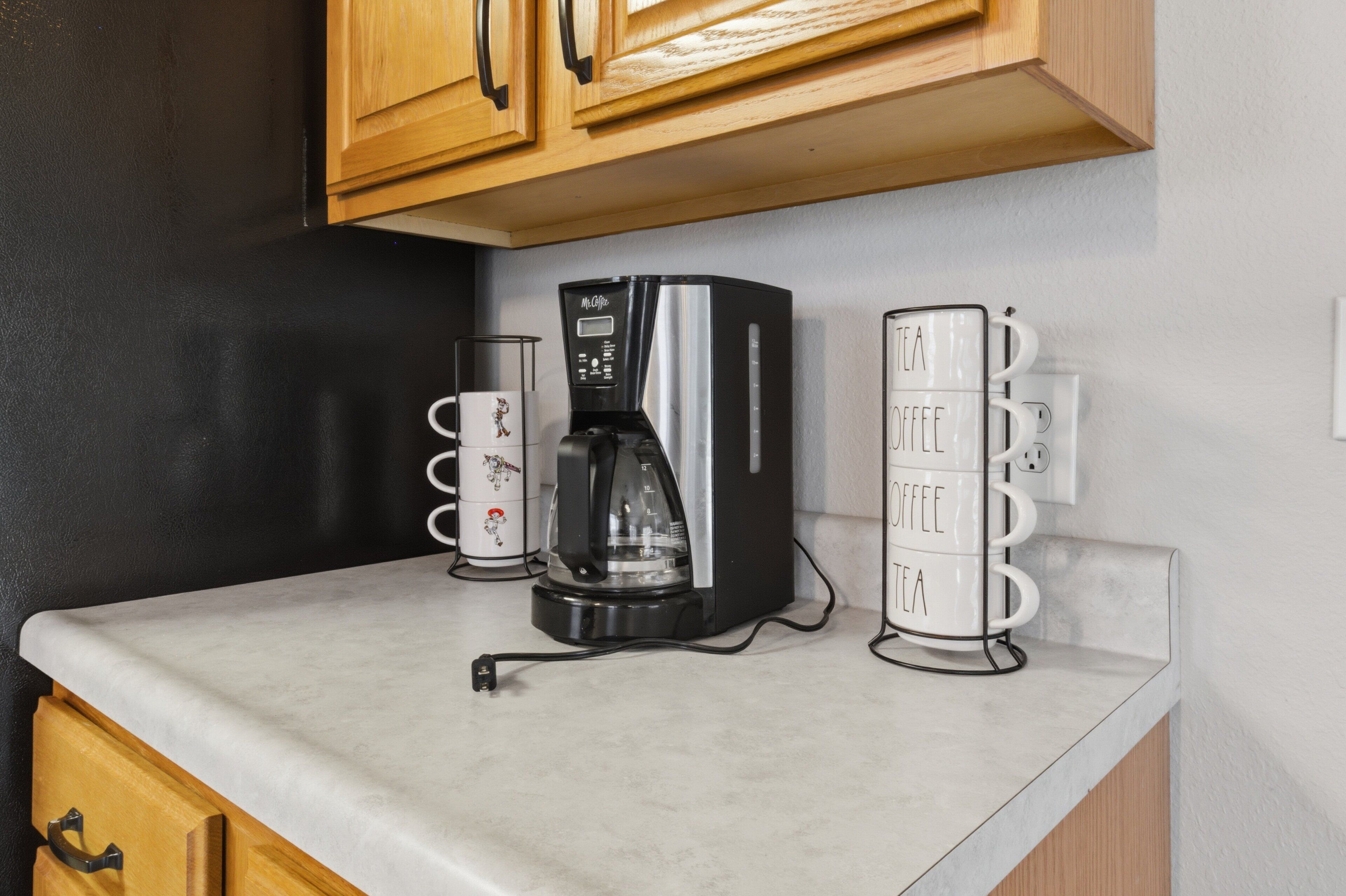 A kitchen with a coffee maker, a stack of tea and coffee mugs, and wooden cabinets.