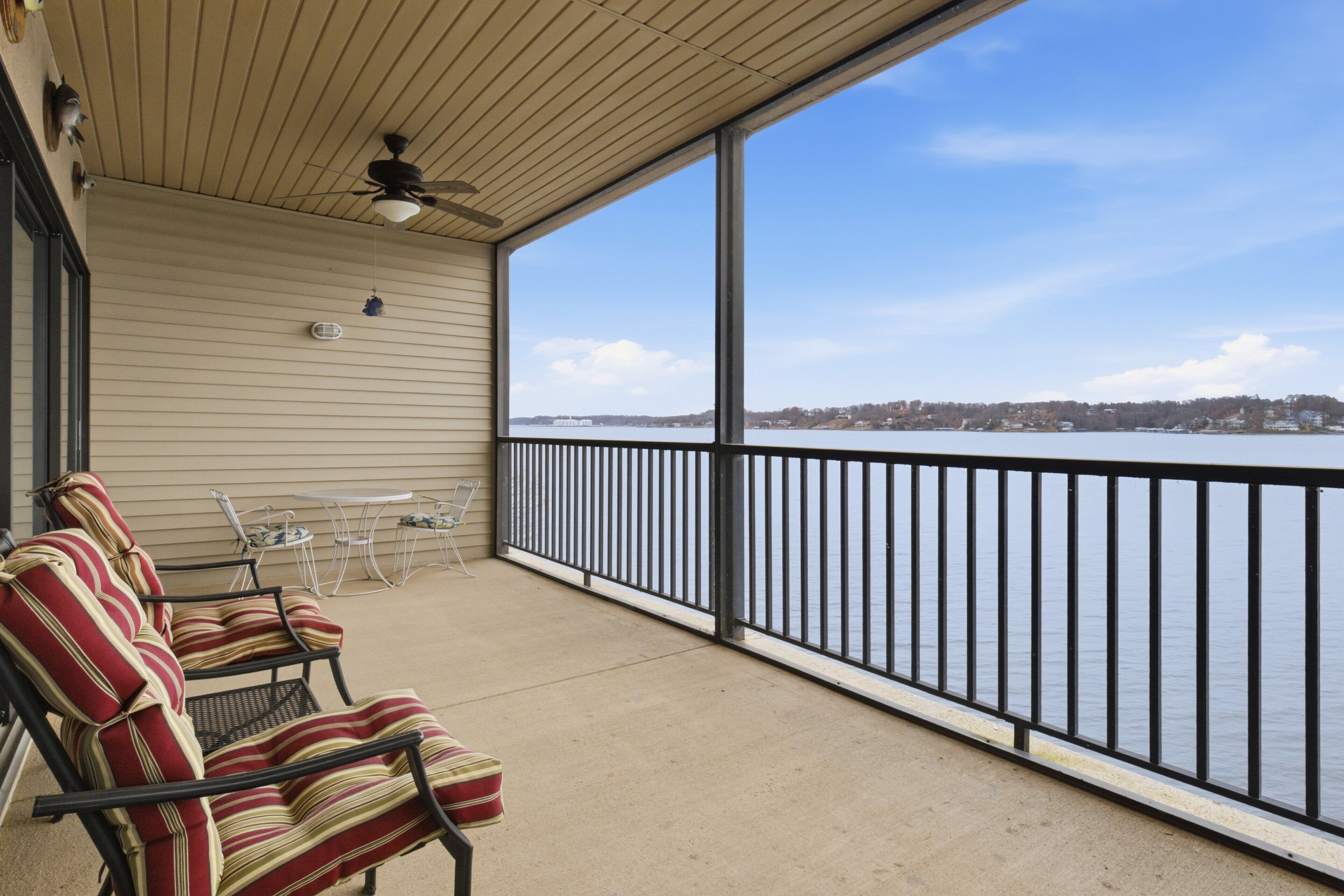 A covered balcony with striped chairs and a table, overlooking a body of water with a distant shoreline.