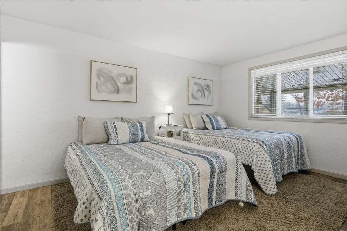 A bedroom with two beds, each with patterned bedding, a nightstand with a lamp, and two abstract paintings on the wall.