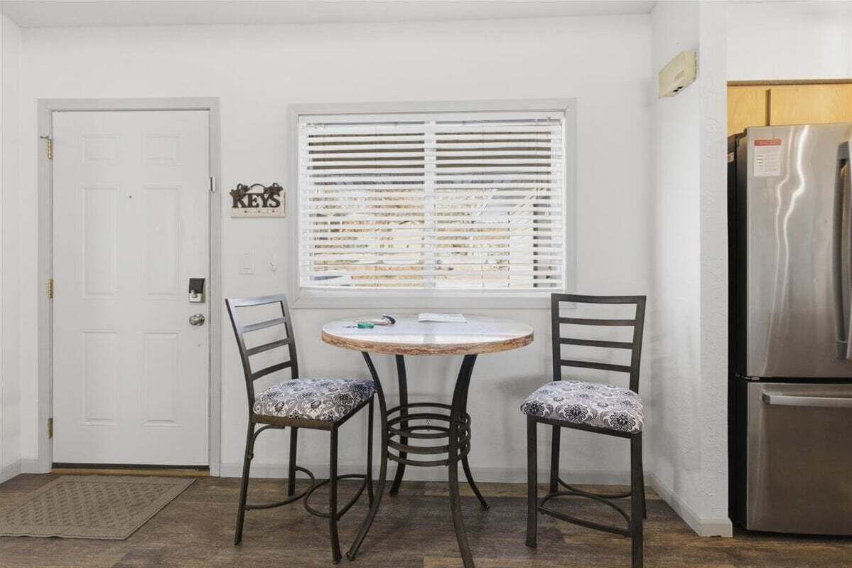 Een kleine keuken met een witte deur, een raam met jaloezieën, een roestvrijstalen koelkast en twee gestreepe stoelen rond een ronde tafel.