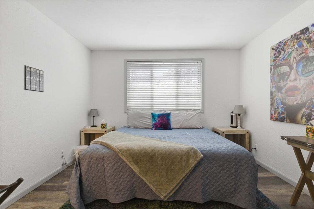 Quarto com cama, duas mesas de cabeceira, janela com persianas, uma obra de arte na parede e um tapete pequeno.