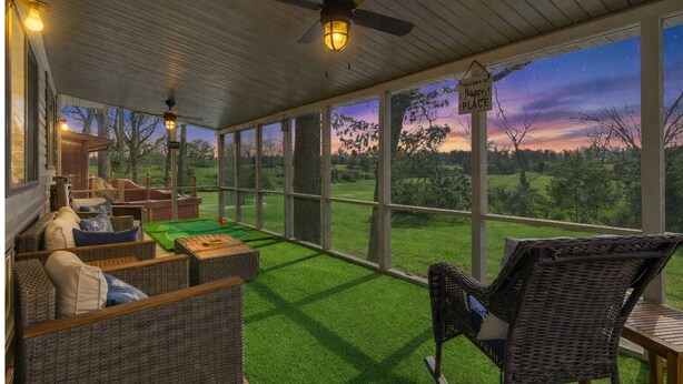 Een overdekt terras met rieten meubilair, een plafondventilator en uitzicht op een zonsondergang over een grasachtig landschap.