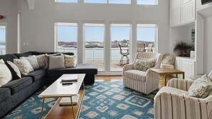 Ein geräumiges Wohnzimmer mit einer großen Eckcouch, einem gestreiften Sessel und einem Couchtisch. Mehrere Fenster bieten Blick auf den Strand und die Skyline.