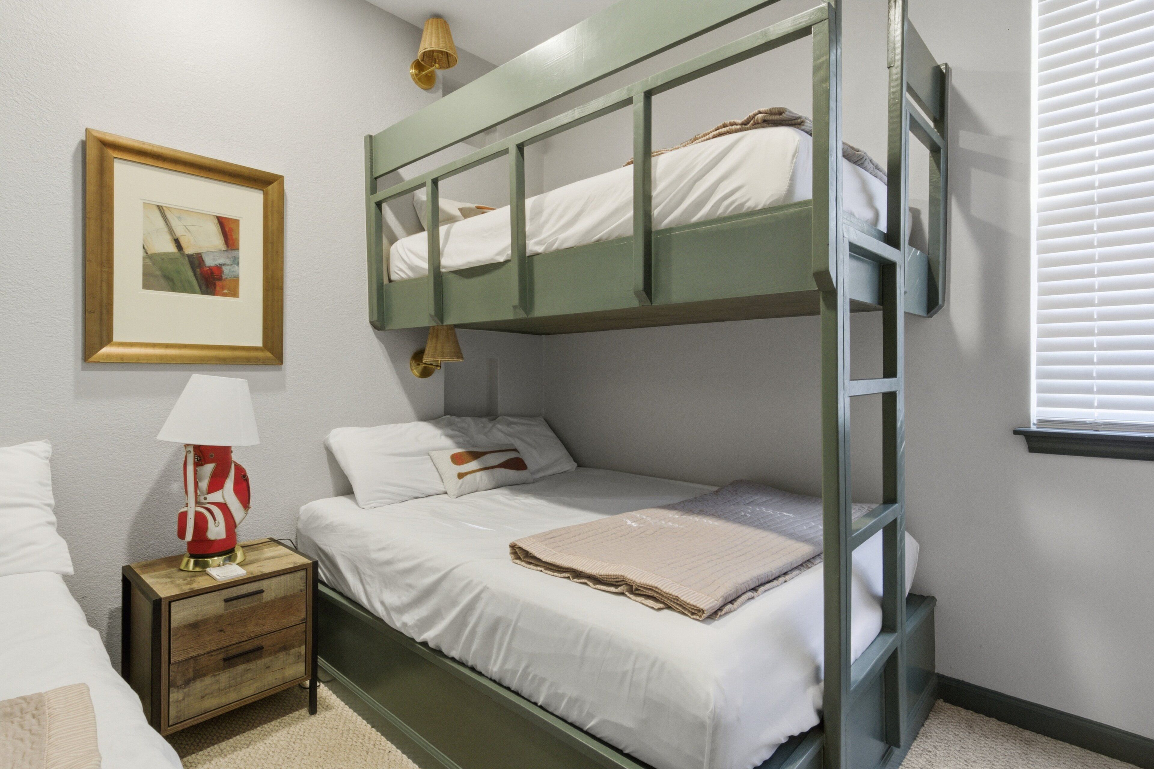 A bunk bed room with a nightstand, a framed picture, and a window with blinds.
