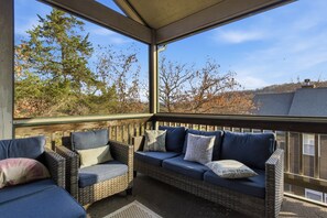 A covered outdoor seating area with wicker furniture and cushions, overlooking a wooded landscape. - Old Kinderhook 2 Decks, Firepit, Kid & Pet Friendly Golf Retreat (Camdenton)