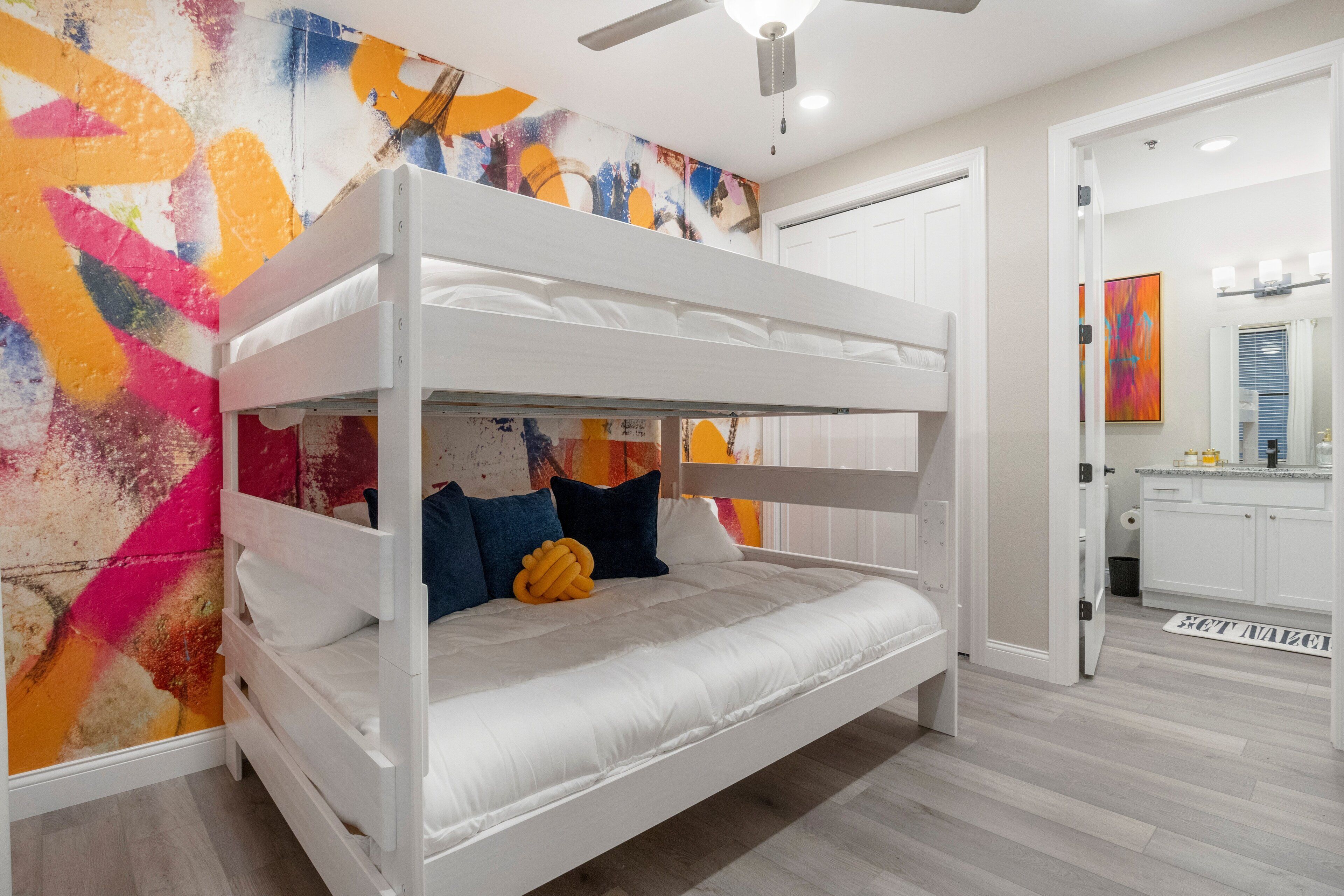 Una cama literera en una habitación con una pared de diseño abstracto y colores vivos, un ventilador de techo y una cocina visible a través de una puerta abierta.