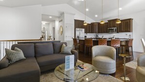 A modern living room with a sofa, armchairs, and a glass coffee table. A kitchen with wooden cabinets and a dining area is visible in the background. - Fairway Retreat (Camdenton)