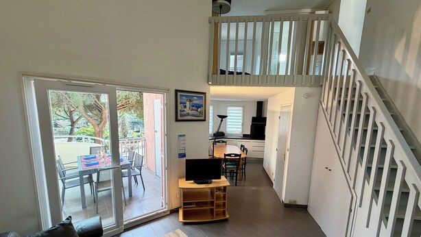 A modern living room with a sofa, a wooden coffee table, and a TV stand. There is a balcony with a table and chairs, and a staircase leading to an upper level. - Bright Duplex in Cavalière – Steps from the Beach (Le Lavandou)