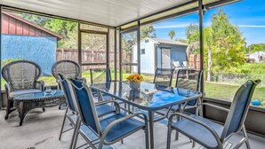 Eine überdachte Terrasse mit Glastisch und -stühlen, ein kleiner Garten und ein blauer Schuppen im Hintergrund.