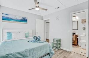 Ein Schlafzimmer mit einem Bett, einem Deckenventilator, einem Strandbild und Blick ins Badezimmer.