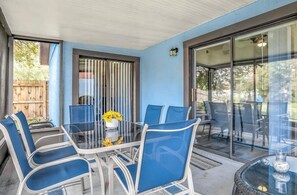 Eine überdachte Terrasse mit Glastisch und blauen Stühlen, einem kleinen Topfpflanzen und einem Korbtisch mit einer Vase.