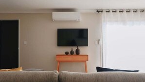 Ein Wohnzimmer mit Flachbildfernseher auf Holzständer, einer Couch und einem Fenster mit durchsichtigen Vorhängen.