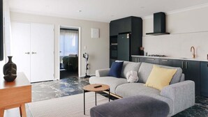 Ein modernes Wohnzimmer mit einem grauen Sofa, einem kleinen runden Tisch und einer Küchenzeile mit dunklen Schränken und einer schwarzen Dunstabzugshaube.