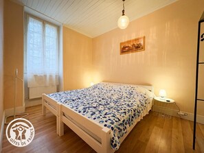 A bedroom with a bed, a window with curtains, a wall-mounted lamp, and a painting. - Gîtes de France® - Patou's Gîte (CATUS)