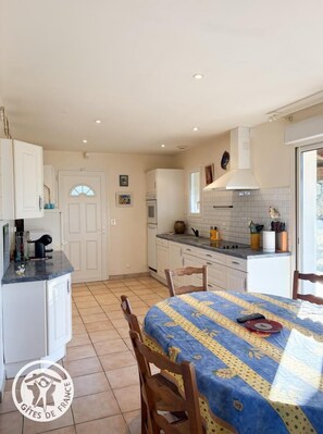 Une cuisine avec des meubles blancs, un dosseret carrelé et un coin repas avec une table recouverte d’une nappe à motifs floraux.