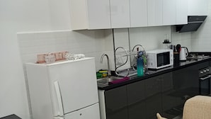 Una cocina compacta con armarios blancos, un refrigerador blanco, un microondas y un lavaplatos con escurreplatos. En primer plano, un sofá color beige.