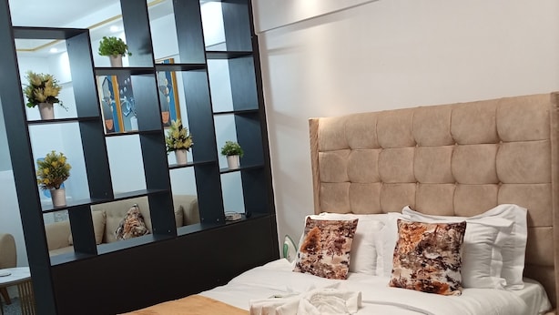 Un dormitorio moderno con cabecera capitonada, cama grande con ropa de cama blanca, cojines decorativos y una estantería negra con plantas.