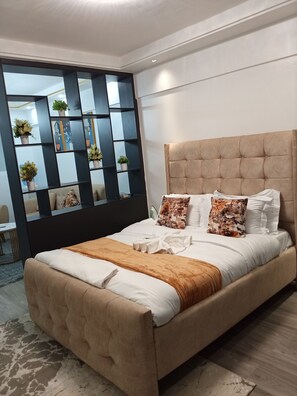 Un dormitorio moderno con cabecera capitonada, cama grande con ropa de cama blanca, cojines decorativos y una estantería negra con plantas.