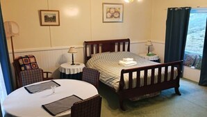 Una habitación acogedora con una cama, una mesa redonda y una silla. Hay dos cuadros en la pared, una lámpara en una mesita de noche y una ventana con cortinas.