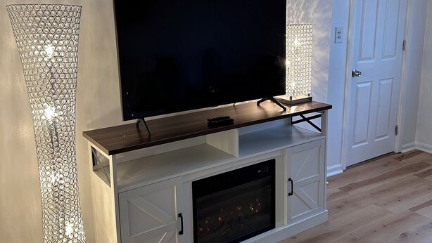 A modern living room with a flat-screen TV on a wooden TV stand, a tall decorative floor lamp, and a white door. - Farmhouse in the Heart of DowntownAuburn Hills (Auburn Hills)