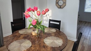 A dining room with a round wooden table, grey upholstered chairs, a vase with pink flowers, and a wall clock. - Farmhouse in the Heart of DowntownAuburn Hills (Auburn Hills)